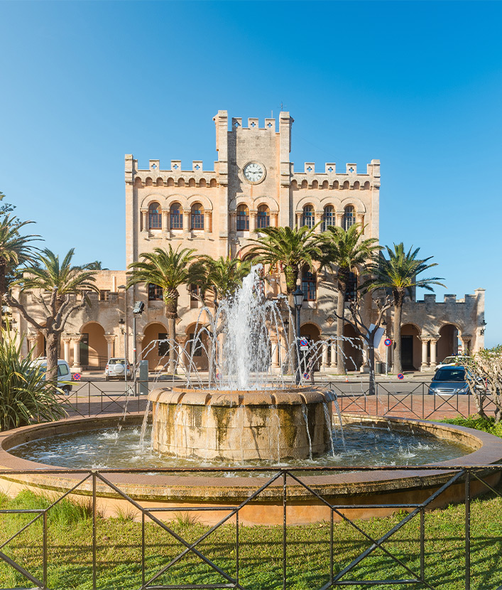Fountain in a park with town hall building in background in Ciutadella on sunny summer day, Menorca island, Spain