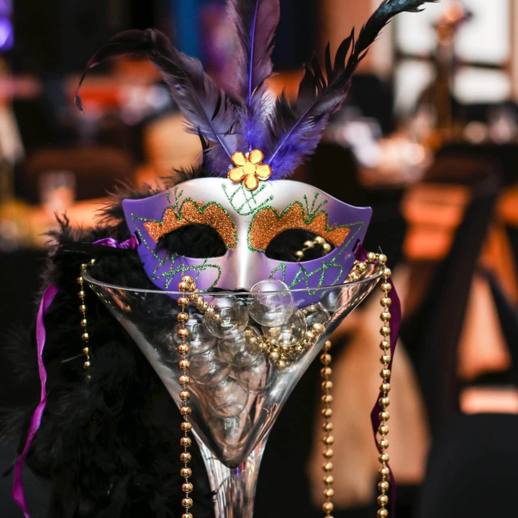 A Mardi Gras mask inside a cocktail glass, with purple feathers